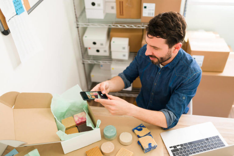 Maumbury Design: A man wearing a blue denim shirt sits at a table taking a photo with his smartphone of a partially open box containing various handmade soaps and items, with cardboard boxes in the background and a laptop on the table.