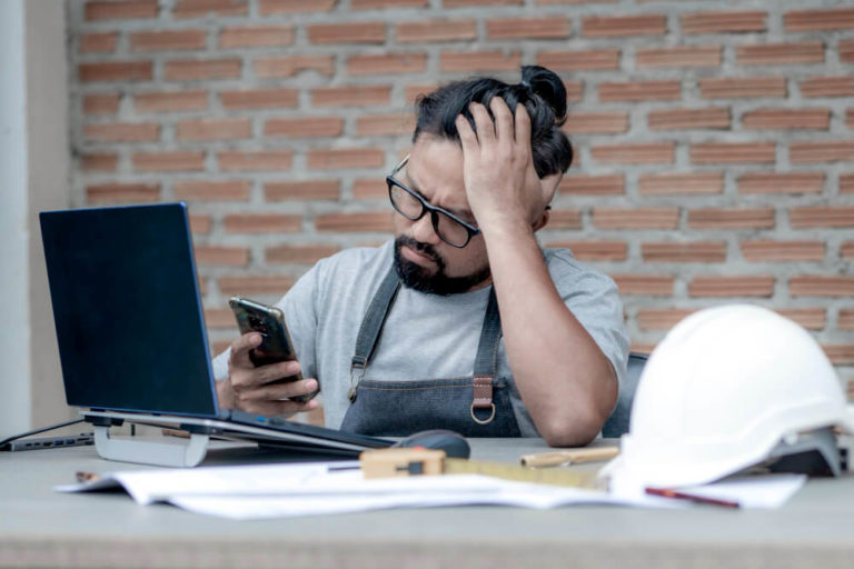 Maumbury Design: A man with a beard and glasses looks stressed while sitting at a desk, holding a phone in one hand and running his other hand through his hair, with a laptop, construction materials, and a hard hat on the table, against a backdrop of brick walls.